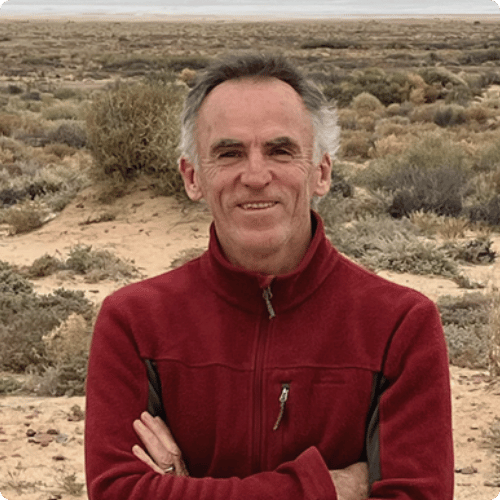 Richard stands with crossed arms in a red jacket with short grey hair, he is standing amongst dry scrubland