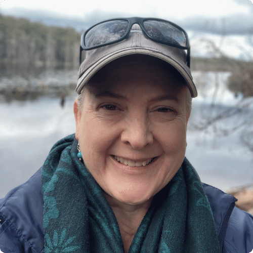 Kylie (she/her) is standing in front of a wetlands. She is wearing a hat, sunglasses and a scarf.