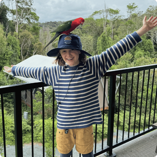 12 year-old Luca (he/him) is standing on a balcony with his hands outstretch. On his hat sits a King Parrot bird.