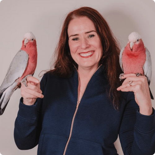 Grainne is holding her pet galahs, one in each hand, and smiling at the camera