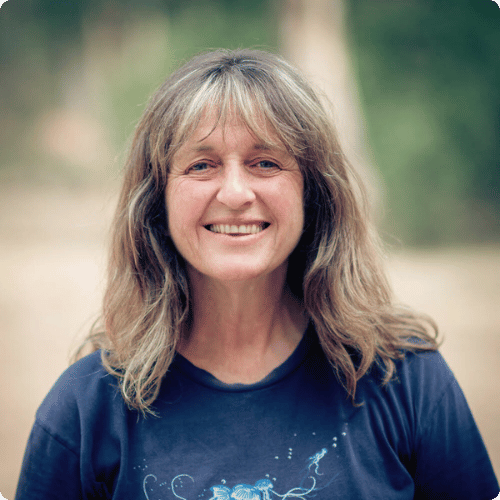 Carol has long blonde hair, she wears a blue t-shirt and smiles at the camera