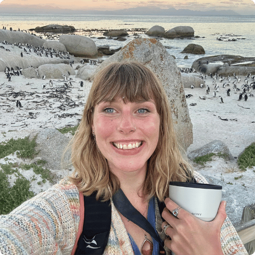 Mia is in South Africa standing in front of penguins. She's holding a camera lens, wearing a backpack and smiling at the camera