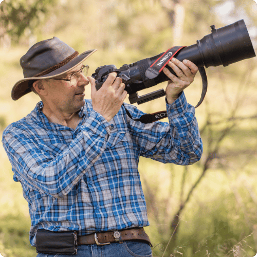 Glenn (he/him) is standing in the bush. He is wearing a hat and plaid shirt. He is looking through a giant wildlife camera lens