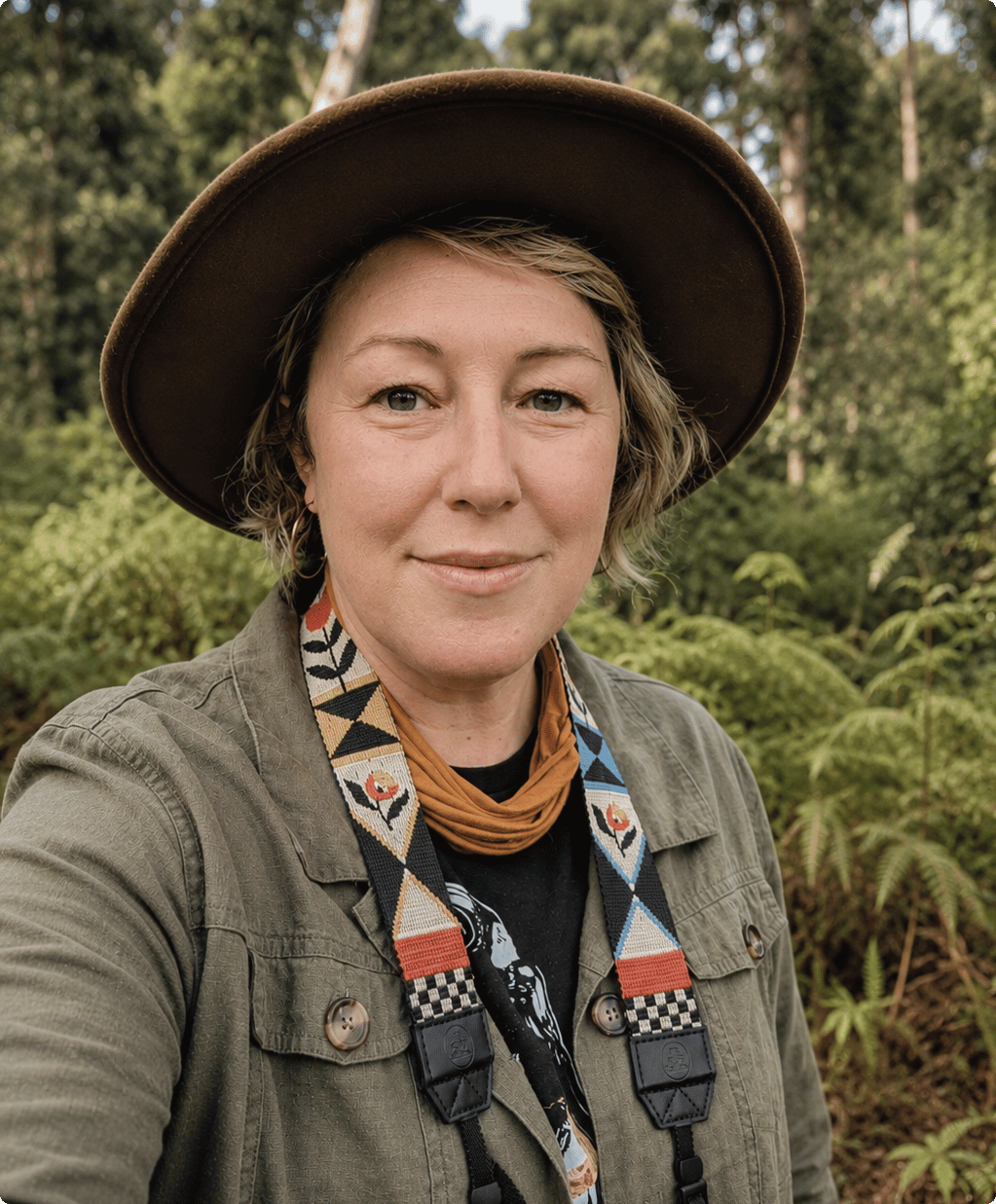 Kirsty is wearing a hat, a jacket, a pair of binoculars and smiling at the camera. She is standing in the bush.