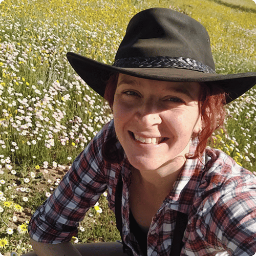Holly is smiling at the camera wearing a wide brimmed hat and sitting in a field of small flowers