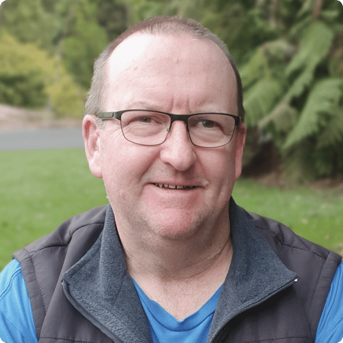 Close up of Anthony smiling at the camera with glasses on and a vest with a greenery in the background