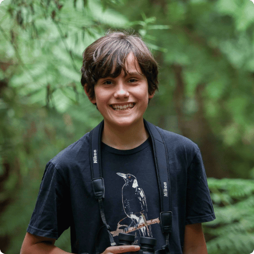 Coen (he/him), a 13-year old birdwatcher, is standing in a forest wearing a magpie tshirt