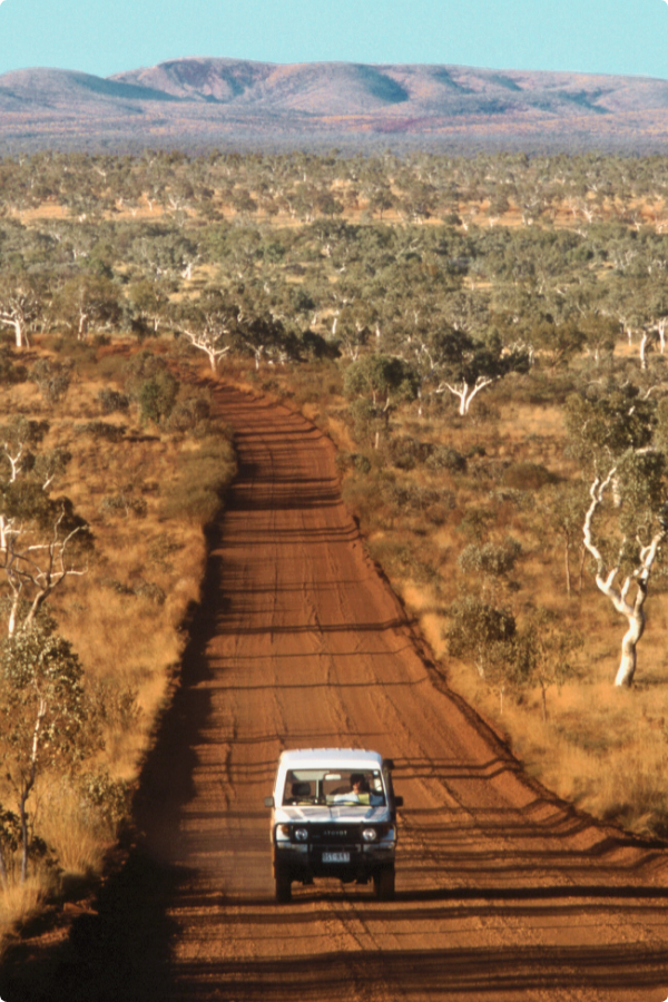 A white 4WD car is driving along a red dirt road with mountains in the background