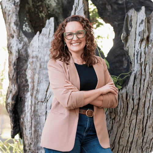 Catherine is wearing a blazer, jeans and glasses. She's standing in front of an old tree stump, arms crossed and smiling at the camera