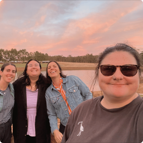 Lucy (she/her) is smiling in front of a dam where the sun is setting. Her friends are leaning in the background