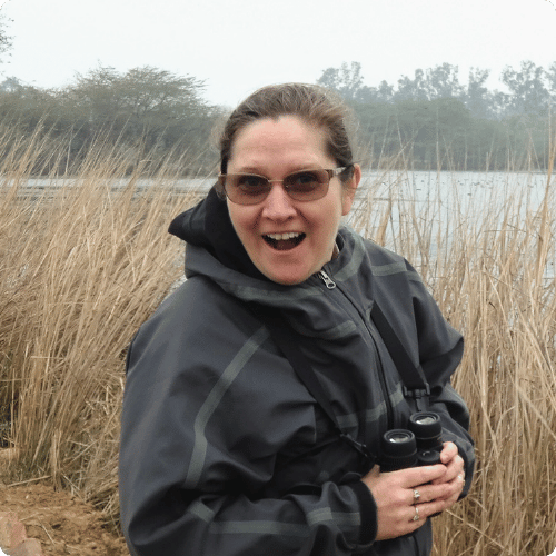 Liz (she/her) is standing in a wetland and has a look of surprise on her face. She is holding binoculars - maybe she's just seen a bird!