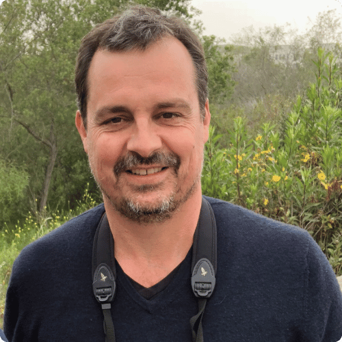 Sean has short grey hair and a beard, he looks smiling at the camera with a strap around his neck