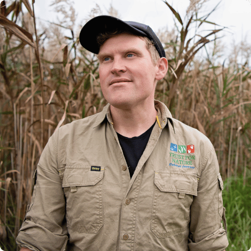 Ben is standing in a wetland, wearing his Trust for Nature hat and shirt