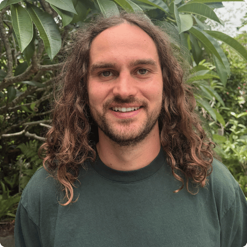 Jonah (he/him) has shoulder length hair and is standing in front of a tree, smiling at the camera