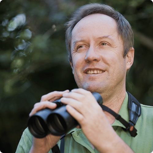 Hugh has short hair and wears a green shirt, he is holding binoculars and looking up into the trees