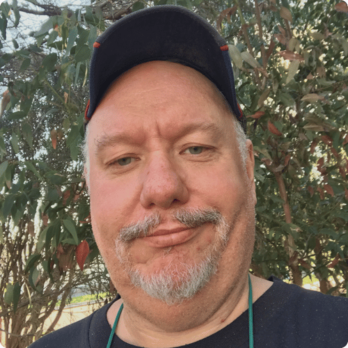 Close up of Grant standing in front of trees, he wears a black cap and has a grey beard & moustche
