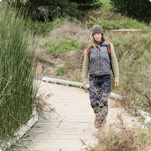 Charlotte is wearing a puffer vest and a beanie and is walking on a boardwalk among nature