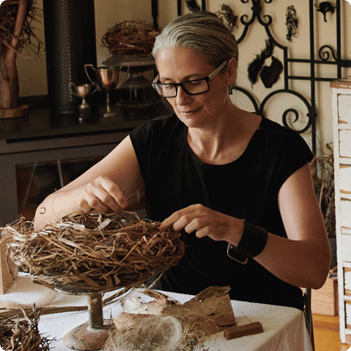 Zora has short grey hair and wears glasses, she is working on a birds nest sculpture in her studio