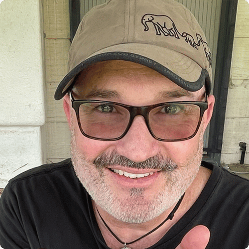 Nicholas wears a cap and sunglasses and is smiling into the camera