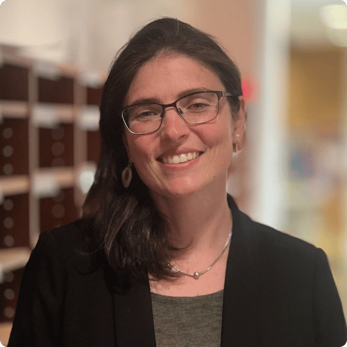 Michelle (she/her) is standing in a library. She is wearing black glasses, a black jacket and is wearing a silver necklace