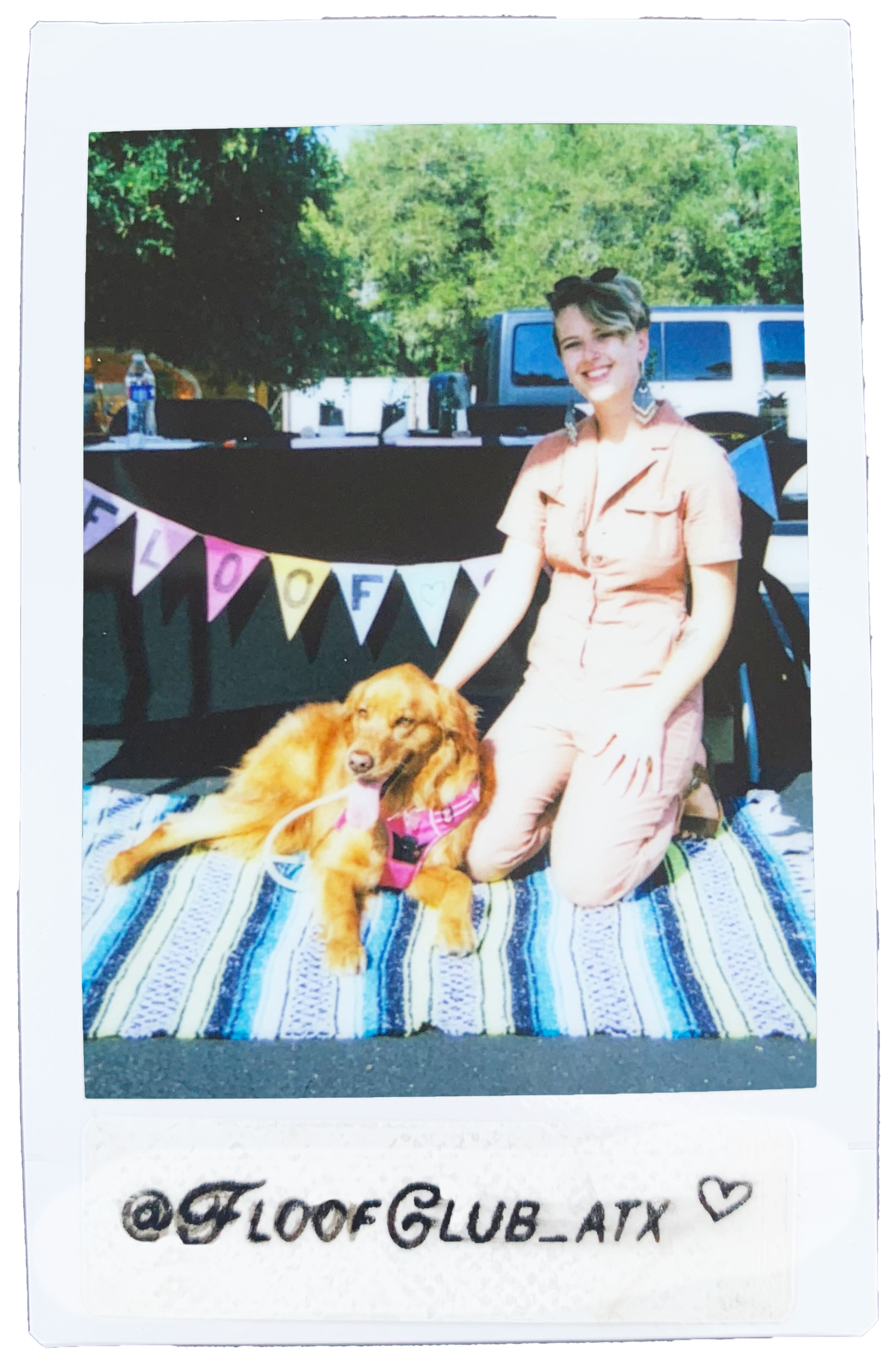 A woman kneeling on a striped blanket outdoors next to a golden retriever dog lying on the blanket. The woman is smiling and wearing pink pajamas. The dog's tongue is out, and it is wearing a pink harness. Behind them is a decorated banner that spells 'FLOOF' with colorful flags. There are trees, cars, and a table with drinks and noise-canceling headphones in the background.