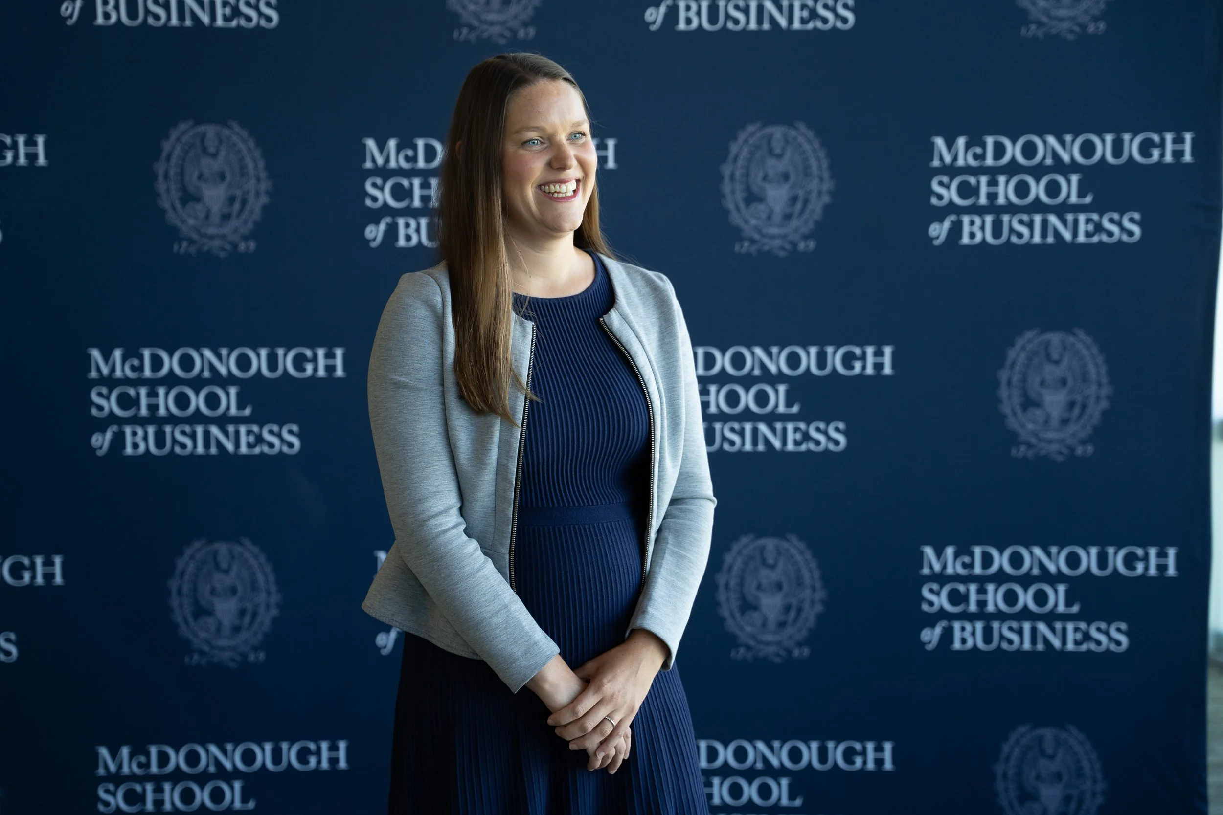 A woman with long brown hair, wearing a gray blazer over a dark blue dress, smiling with her hands clasped in front of her, standing against a blue backdrop with white text that reads 'McDONOUGH SCHOOL of BUSINESS.'