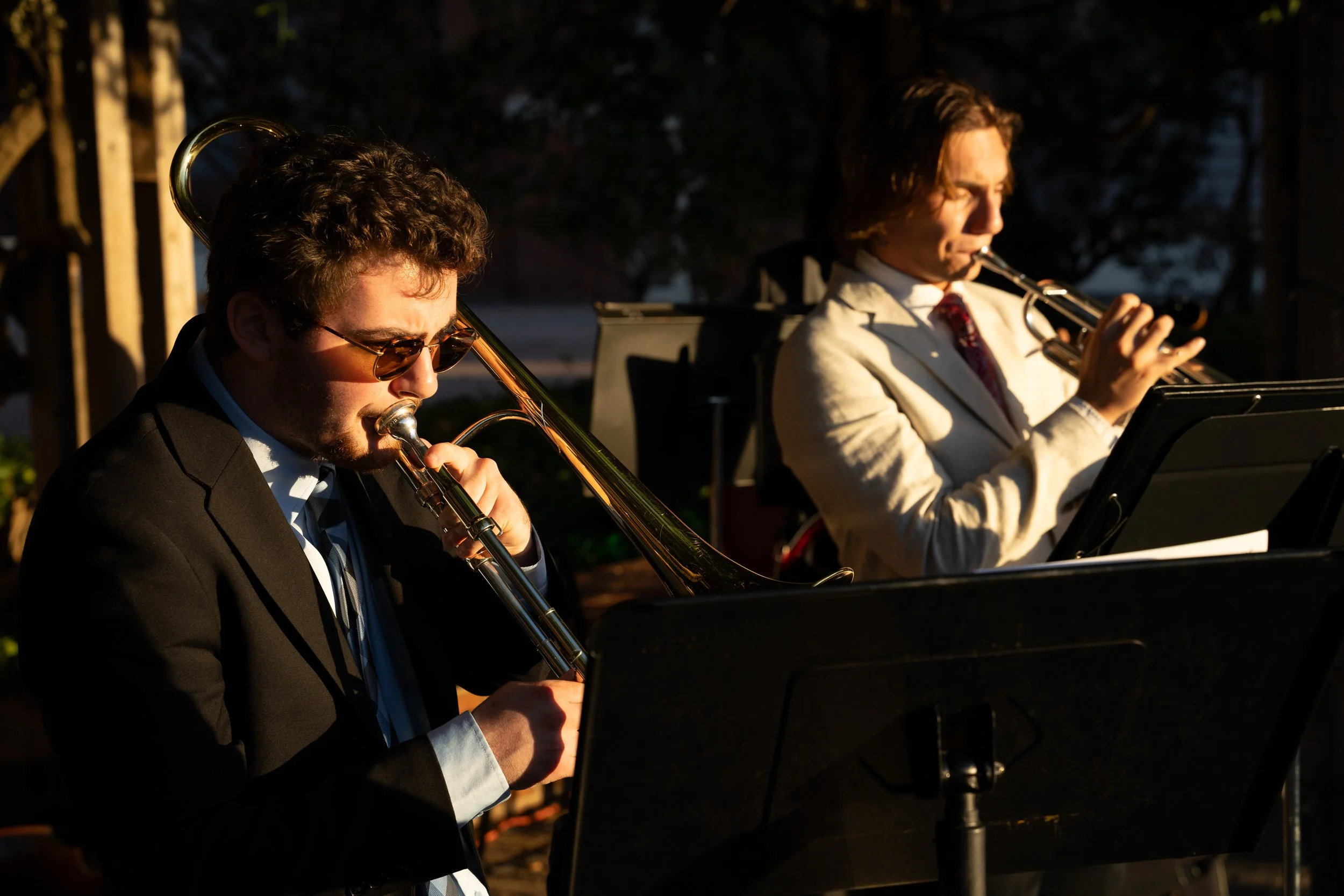 Two musicians playing wind instruments outdoors during sunset, one on a trombone and the other on a trumpet, with music stand and sheet music.