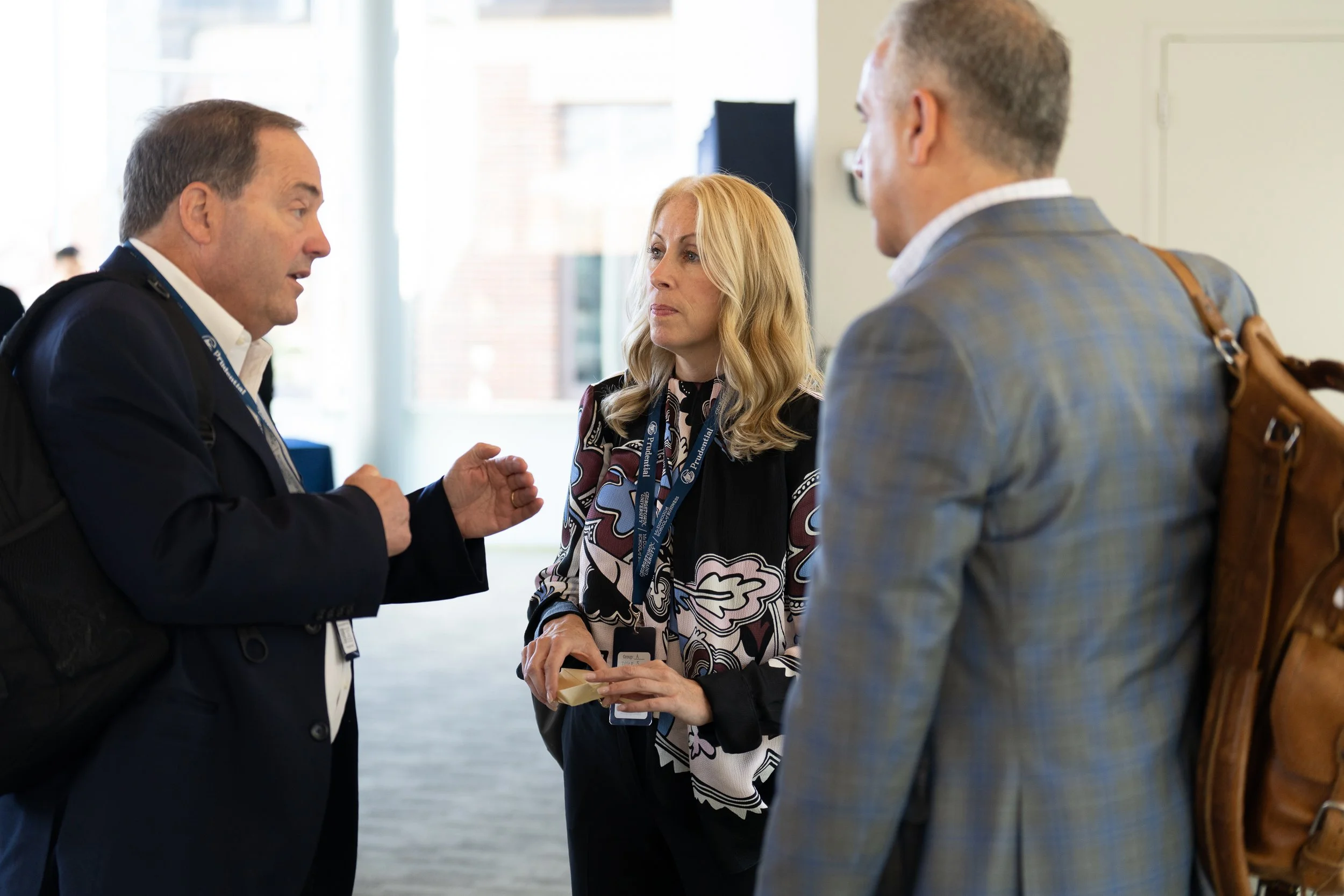 Three people engaged in conversation at a conference or networking event, with one holding a small tray of food or snacks.