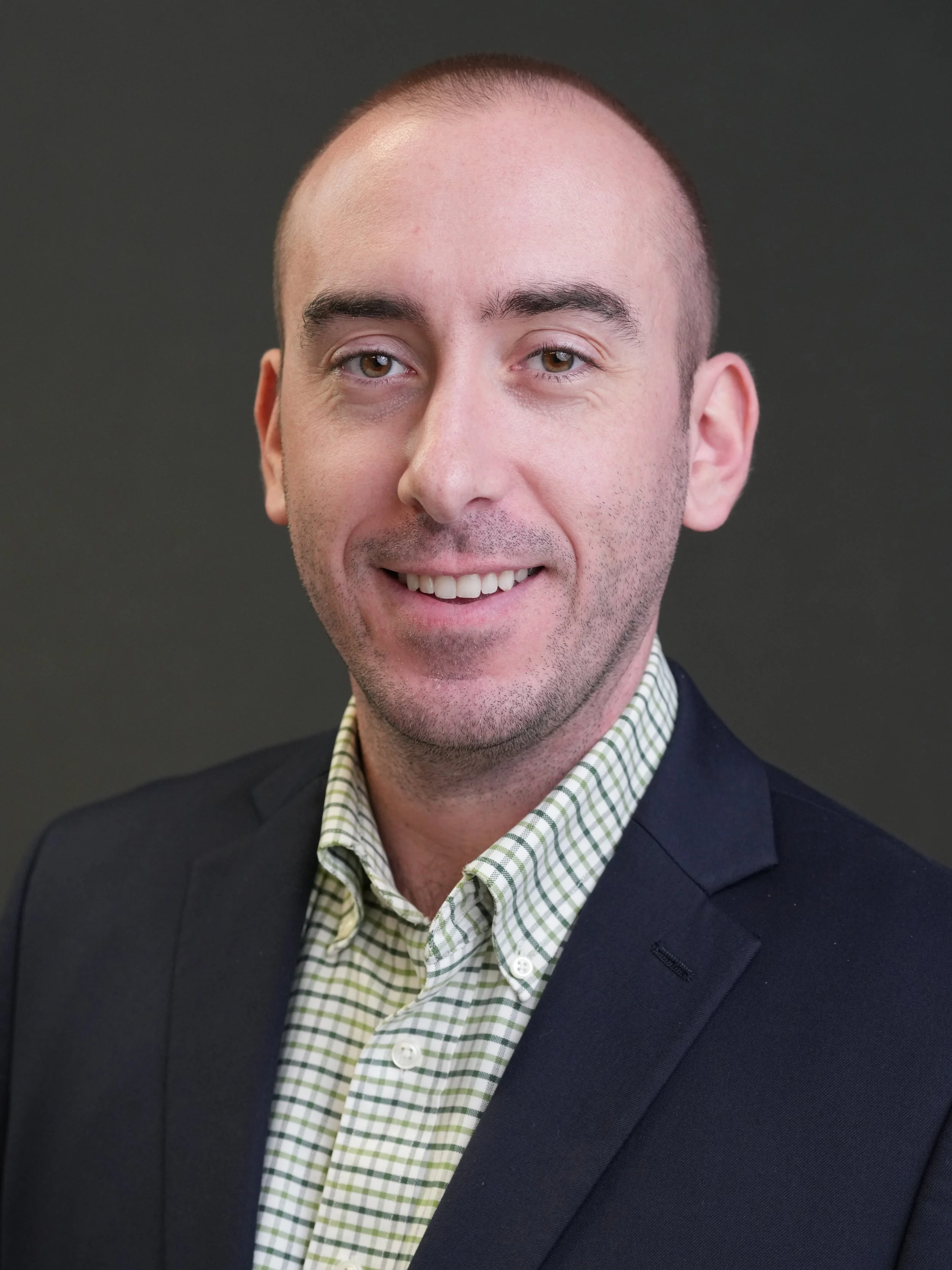 Headshot of a smiling man in a suit jacket and green plaid shirt, against a plain dark background.