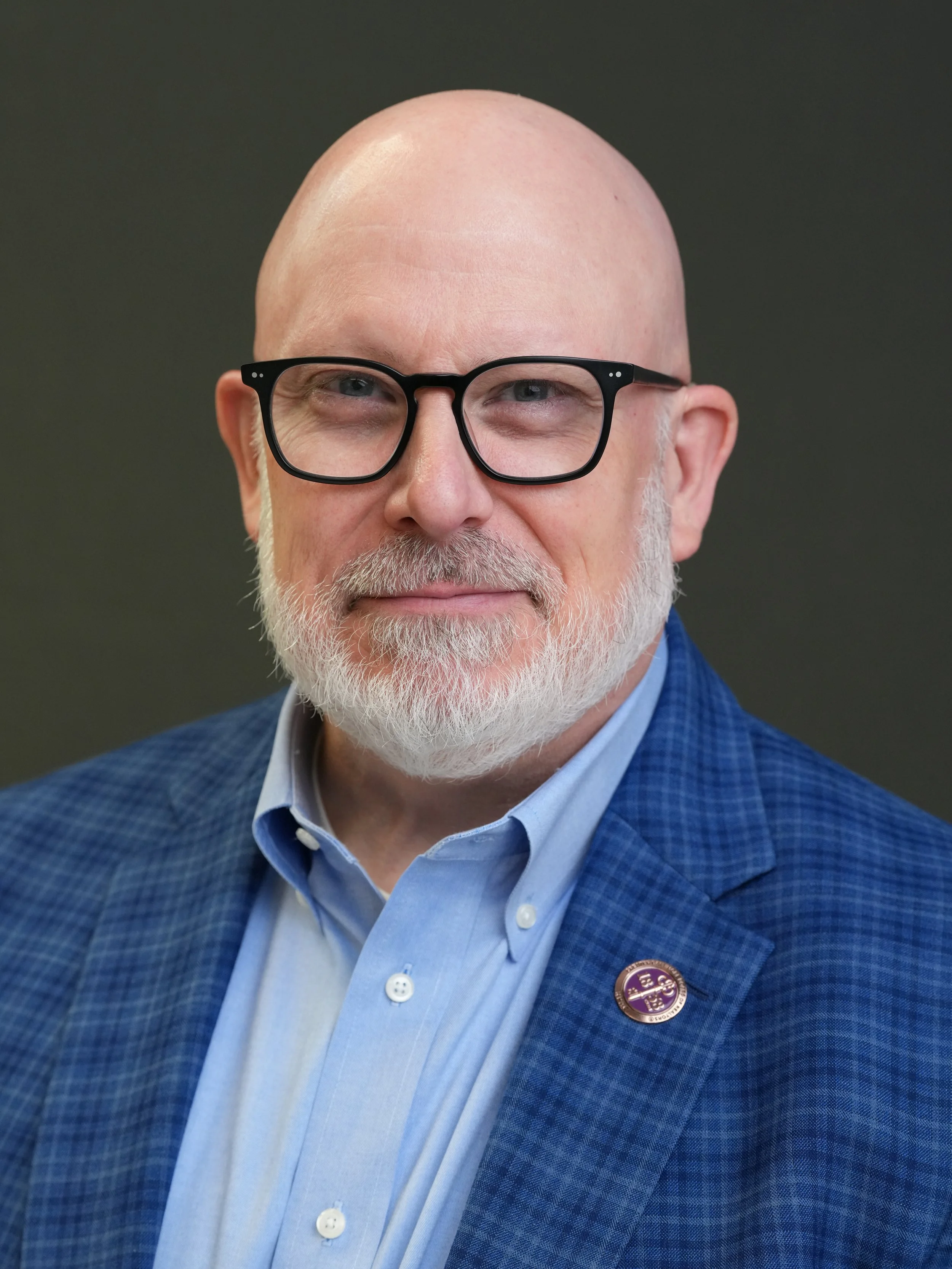 A man with a light beard and glasses, dressed in a blue blazer and a light blue dress shirt, wearing a lapel pin, smiling in front of a dark background.
