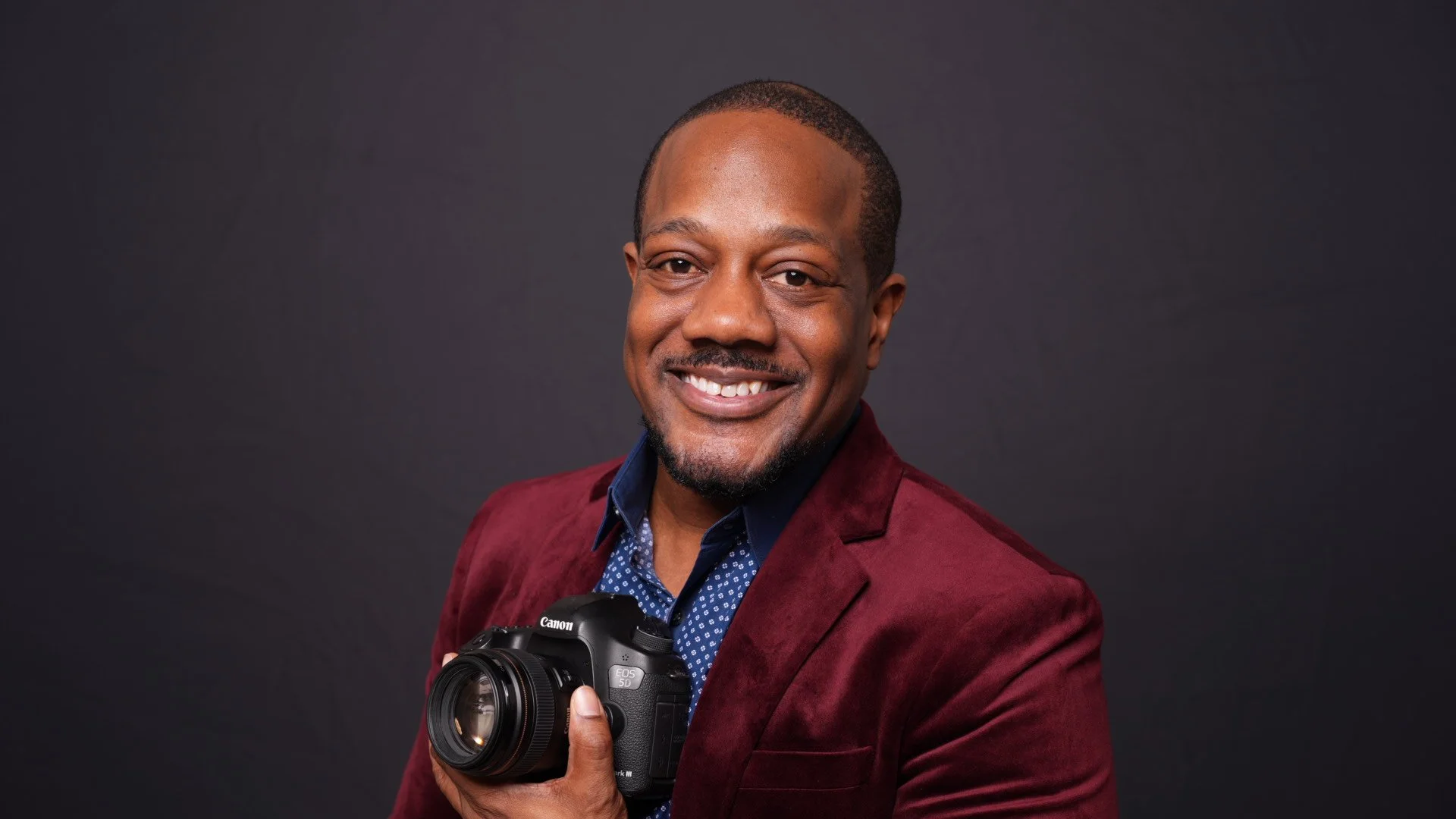 Smiling man holding a camera, wearing a burgundy blazer and a blue shirt with white polka dots, against a dark background.