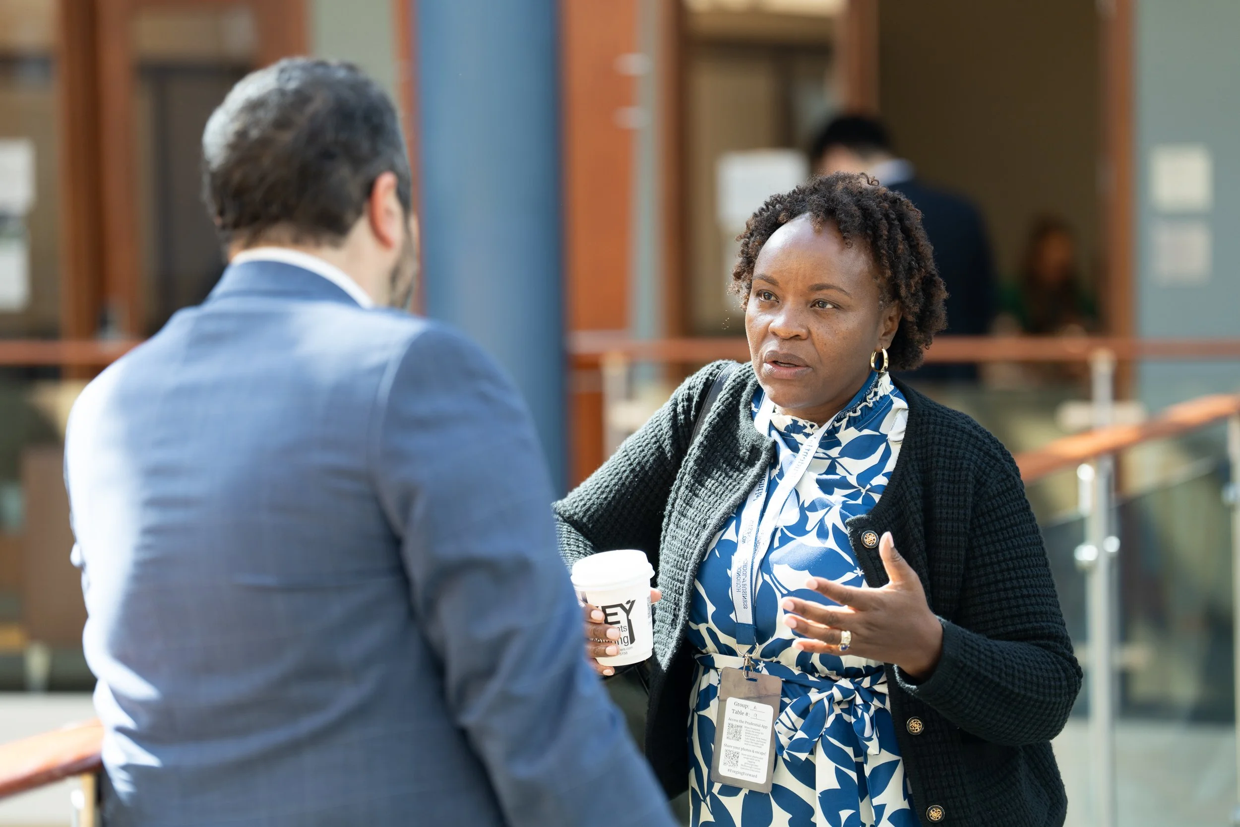A woman with curly hair and a patterned dress is engaged in conversation with a man in a suit at an outdoor event, holding a coffee cup.