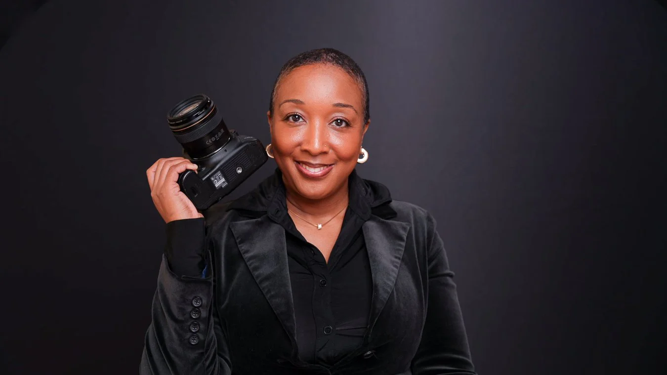 A smiling woman with short hair holding a camera over her shoulder against a dark background