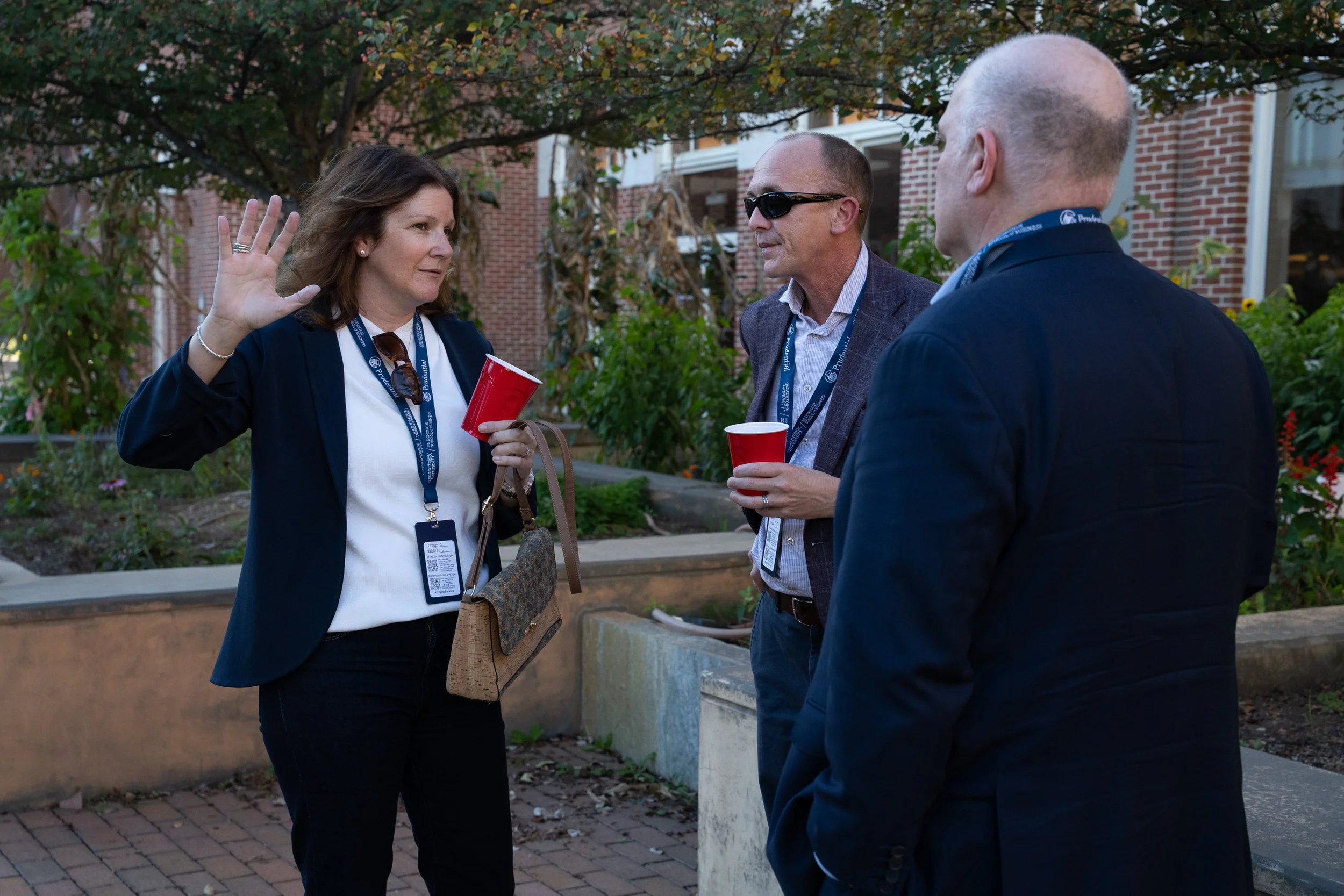 Three people engaged in conversation outdoors near brick buildings and trees, one woman and two men, holding red cups, with name tags on neck lanyards.