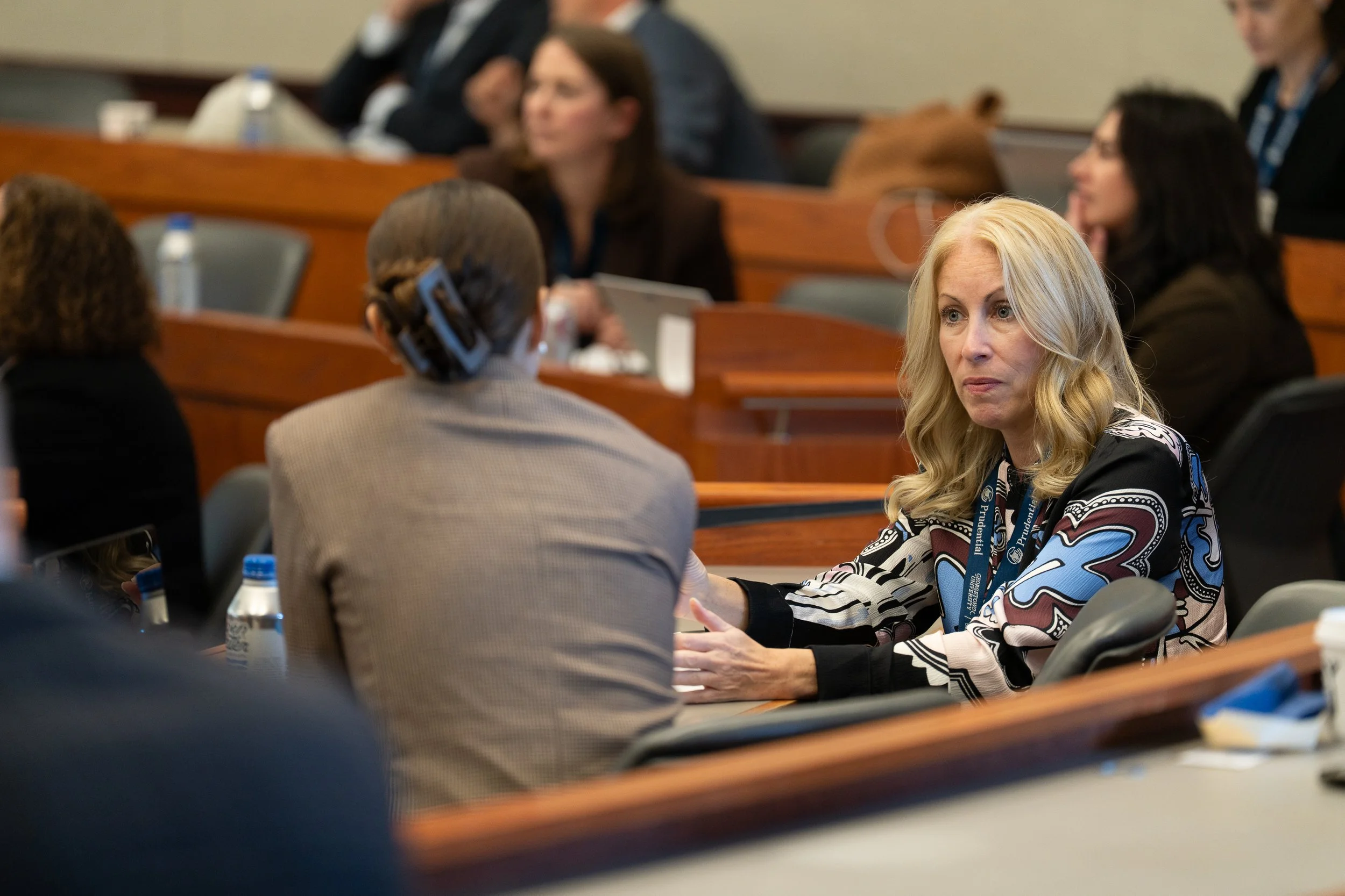 A group of people in a conference room engaged in conversation, including a woman with long blonde hair wearing a patterned blouse and others with their backs turned. There are water bottles and conference badges visible on the tables.