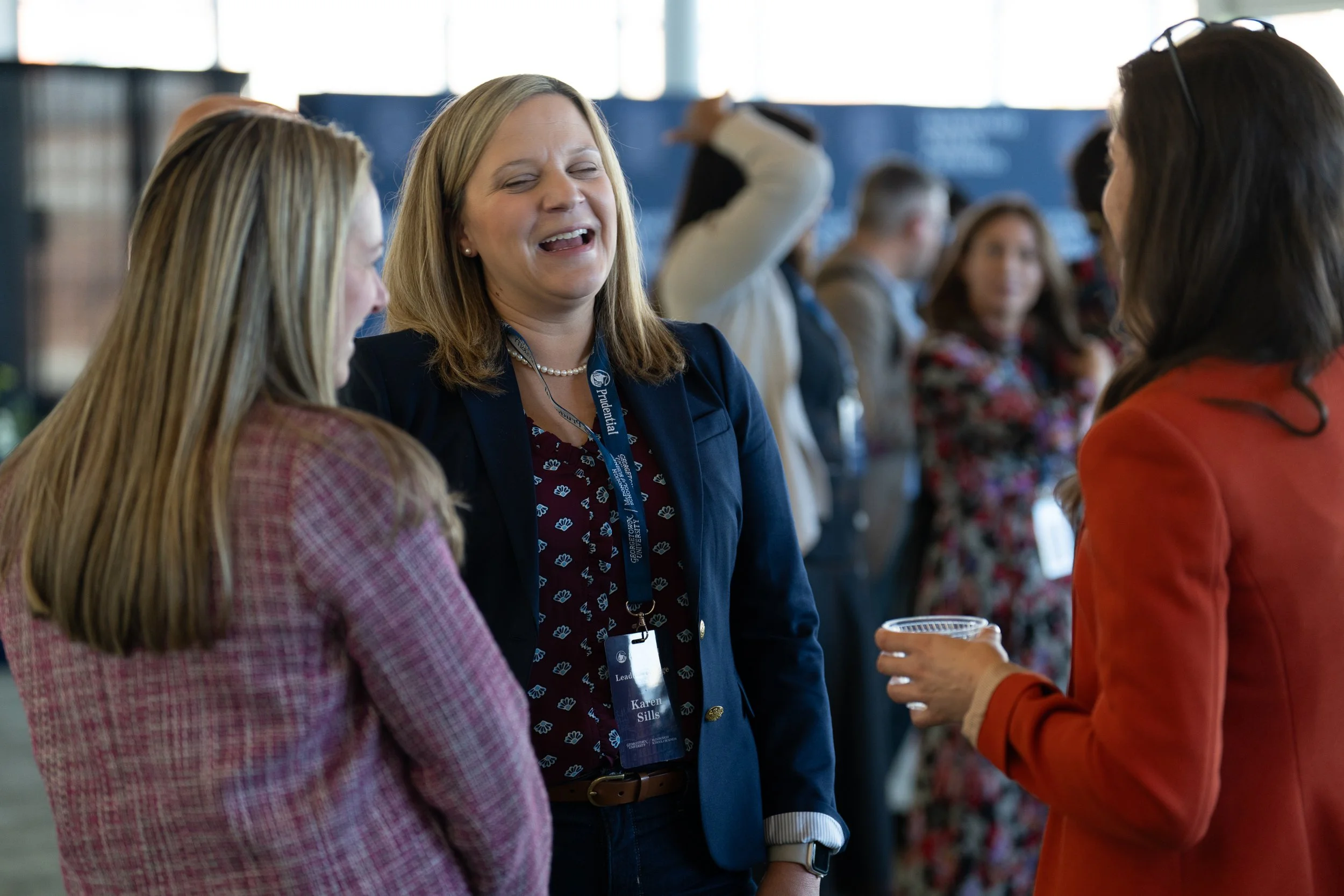 A woman with blonde hair wearing a blue blazer and pearl necklace smiling and talking to another woman with long brown hair wearing an orange blazer, at a professional event or conference.