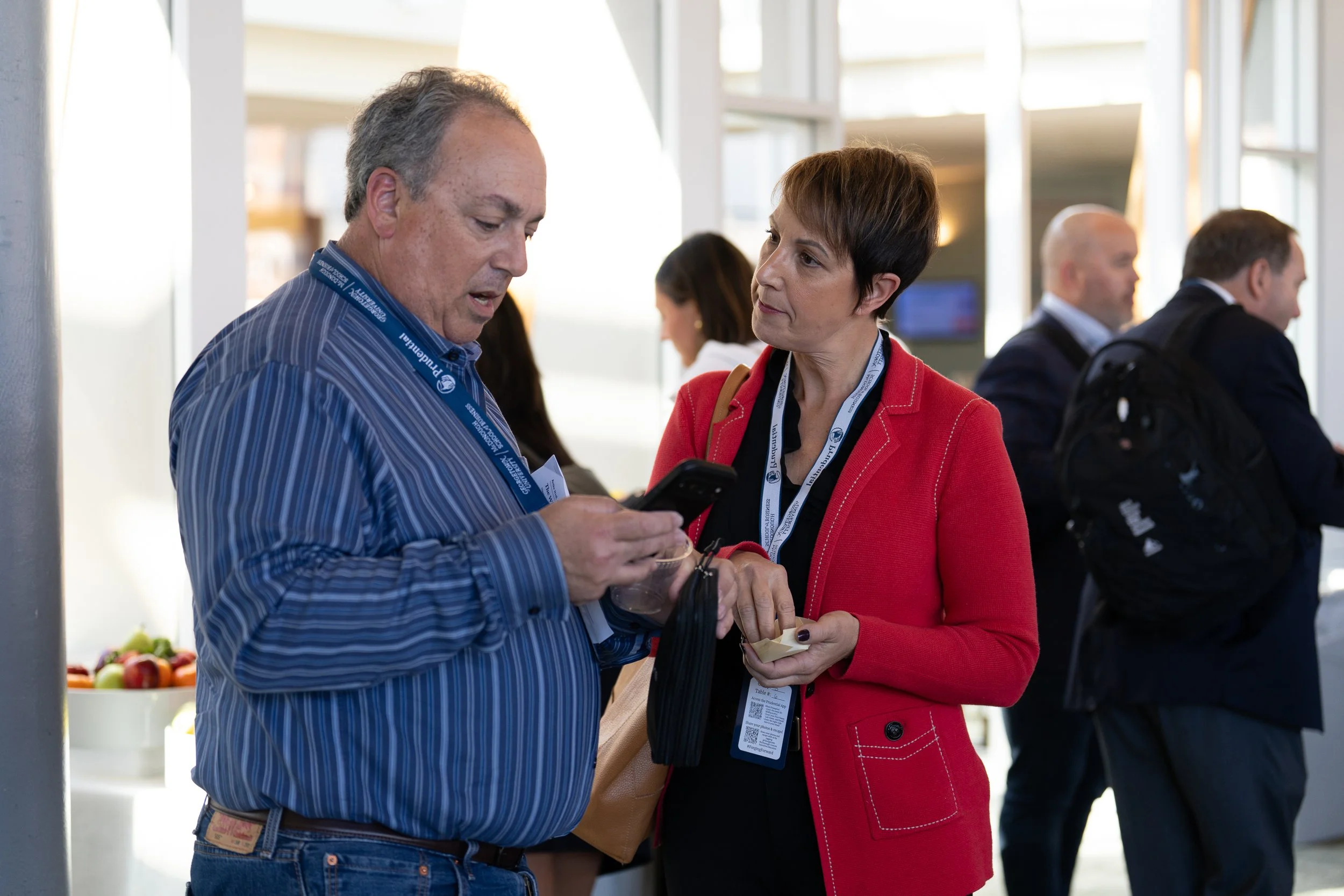 Two people, a man with gray hair wearing a blue striped shirt and a woman with short brown hair wearing a red blazer, are engaged in conversation. They are at a professional event with others in the background, and the man is showing something on his phone to the woman.