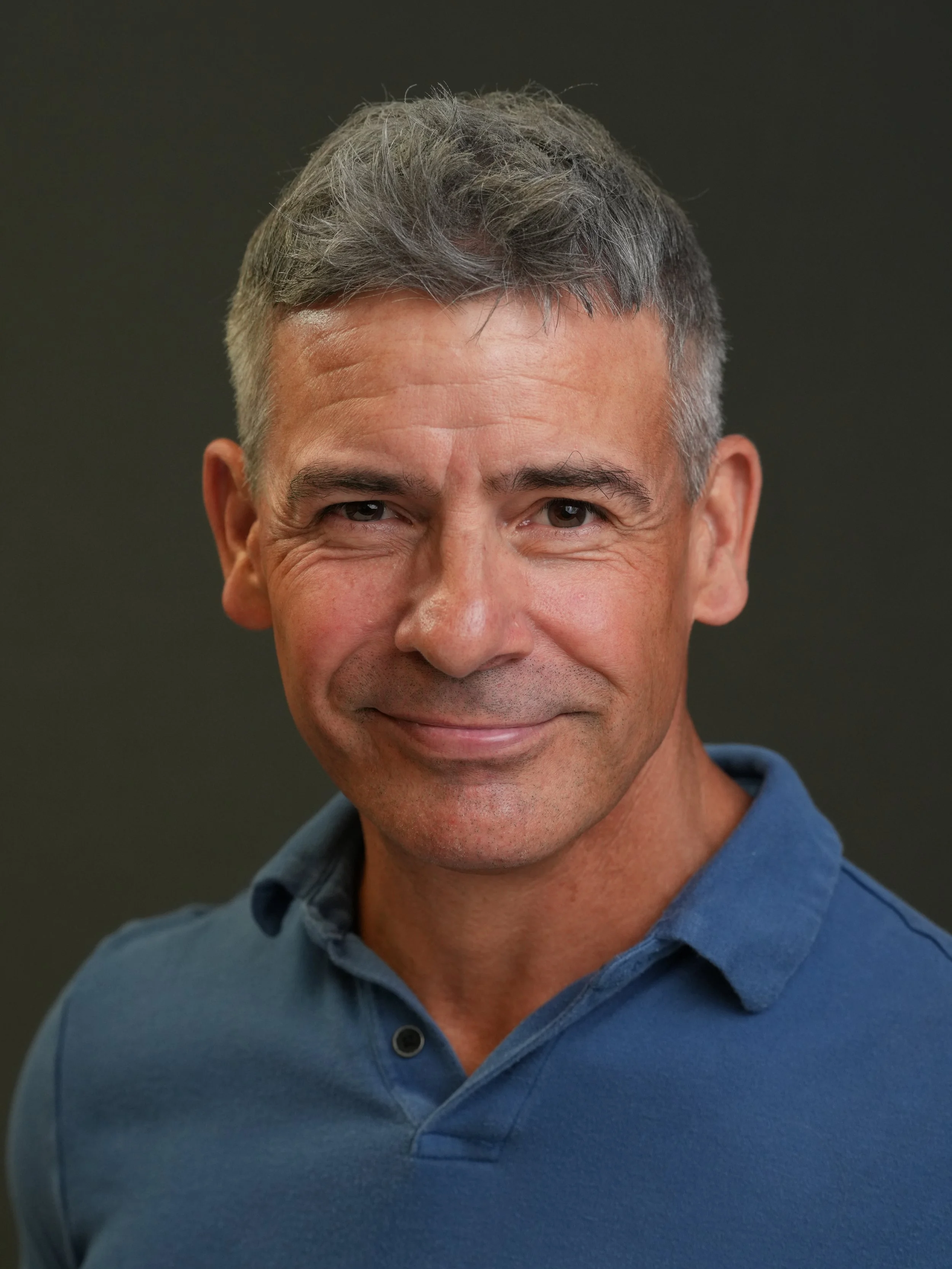 Close-up of a middle-aged man with gray hair wearing a blue collared shirt, smiling slightly, against a blurred dark background.