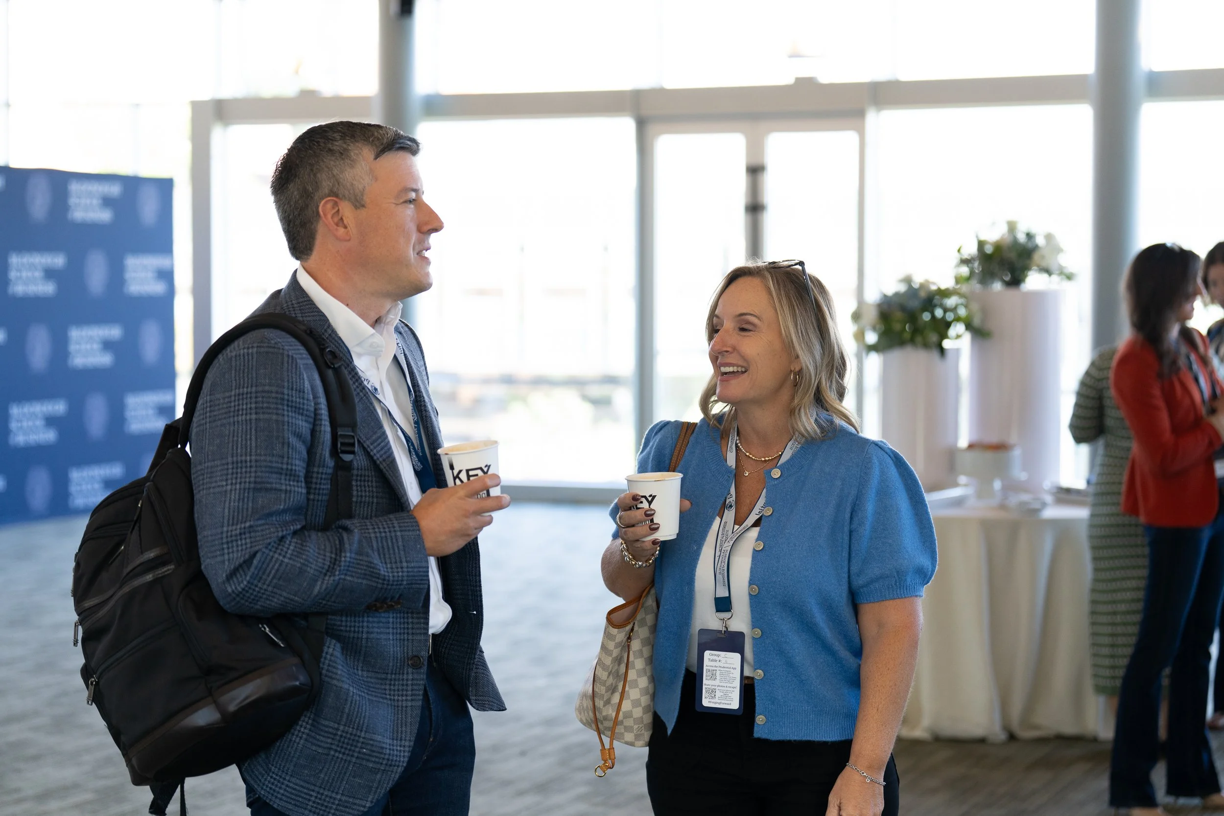 A man and a woman engaging in conversation at a conference or professional event, both holding paper cups, with a backdrop of large windows and other attendees in the background.