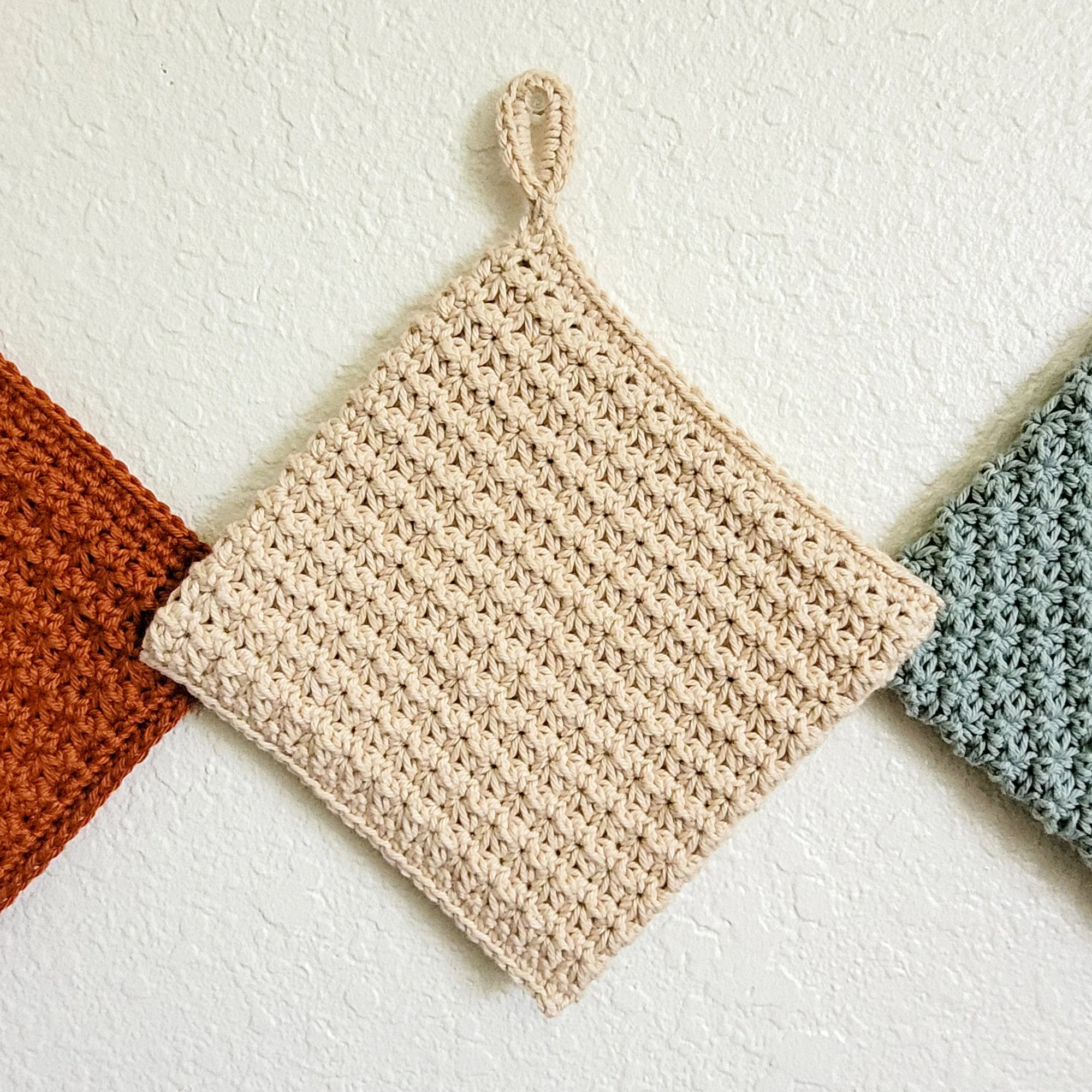Beige Ring-Start Potholder hanging from a wall hook by its crocheted hanging loop. The double-thick Trinity Stitch fabric creates a dense, textured surface.