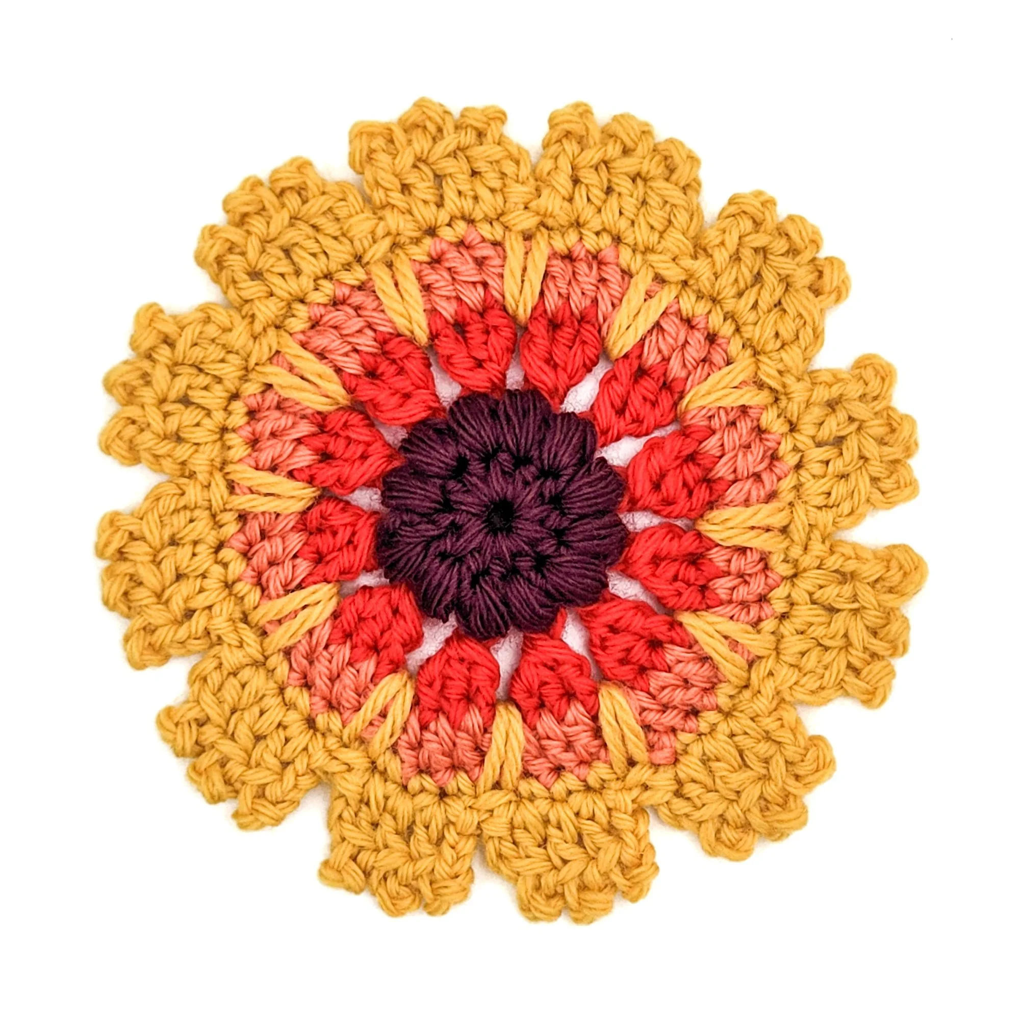 Crochet blanket flower applique on a white background. A puffy, dark purple circle is at the center. Petals radiate outward in shades of bright red, coral orange, and yellow. There are round textural details on the outer edge of the petals.