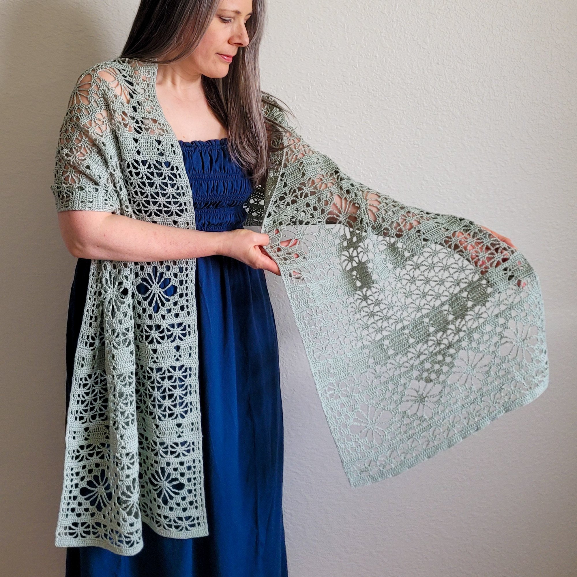 A sage-colored, rectangle-shaped crochet lace wrap is draped over a woman's shoulders. The woman holds out one end to display the lace design.