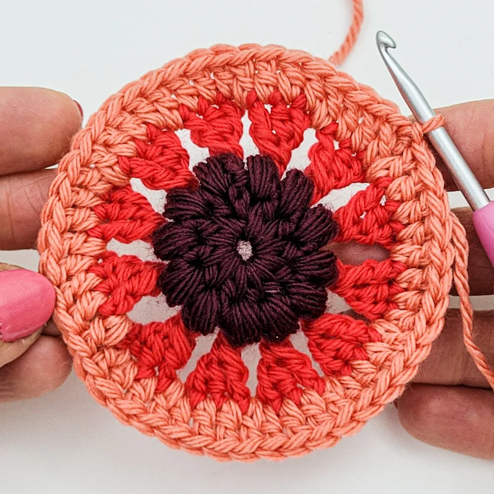 Rnd 5 complete. A crochet circle with red triangles radiating outward from a textured purple center. The outer circle is peach colored.