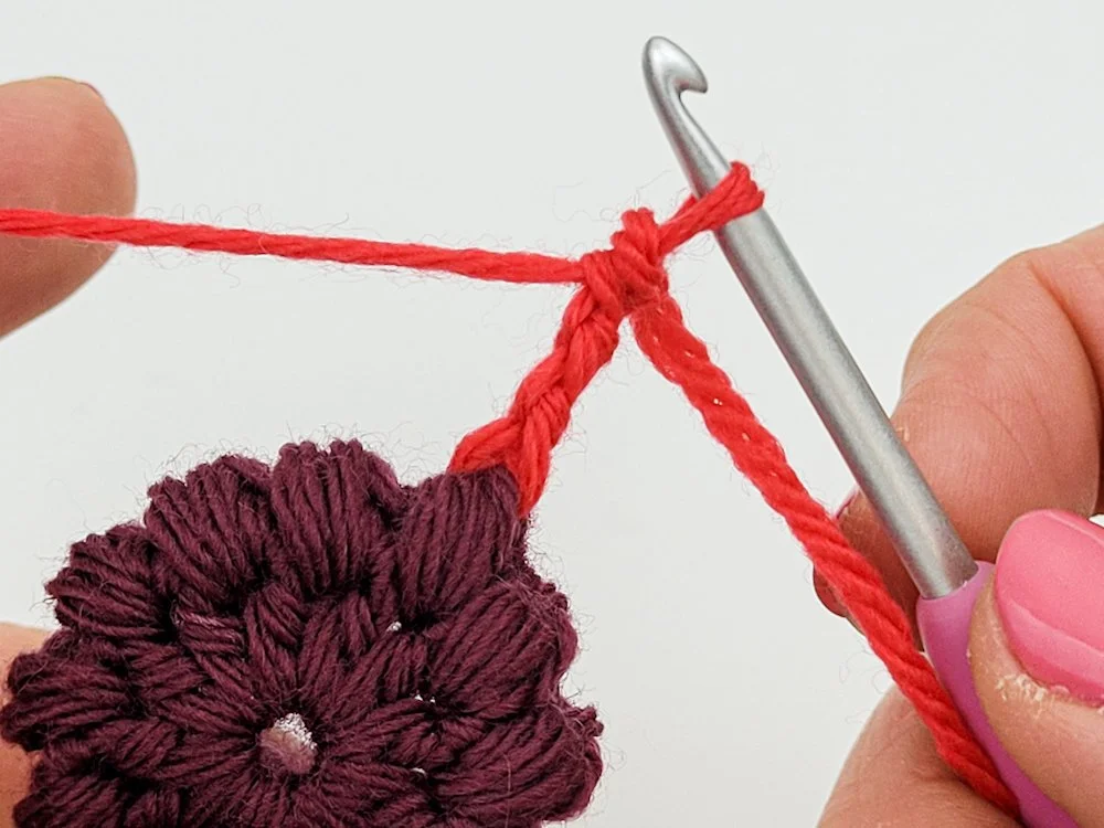 A crochet circle in dark purple yarn. A red yarn has been attached to one of the puff stitches with a standing treble crochet.
