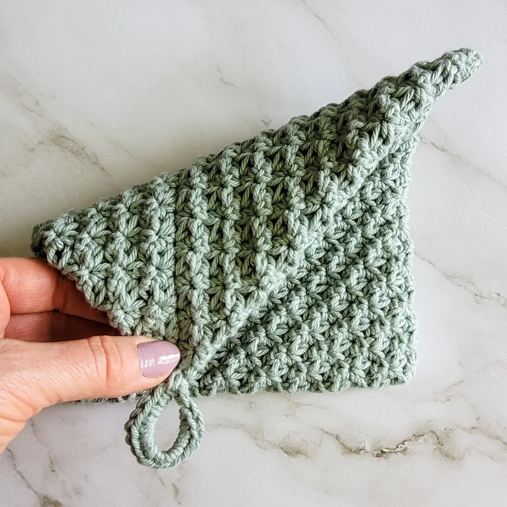 A hand folds the sage green Folded Potholder to show its double-thick construction and distinctive folded design.