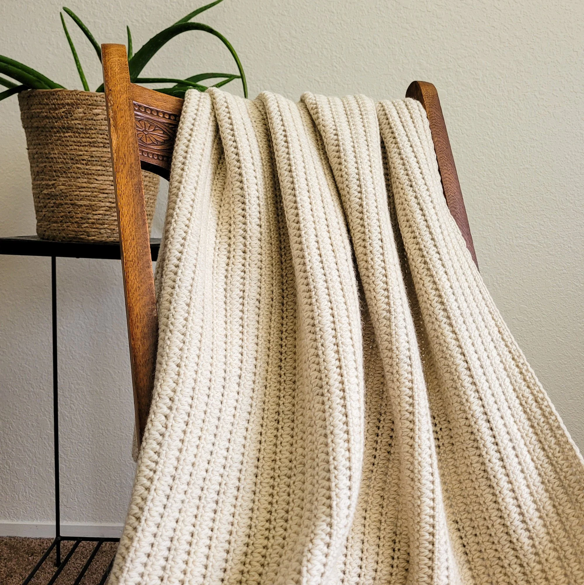 A cream-colored crochet blanket draped over the back of a wooden rocking chair. An interlocking stitch pattern creates a ridged texture along the length of the fabric. A table with an aloe plant in a woven basket is behind the chair.