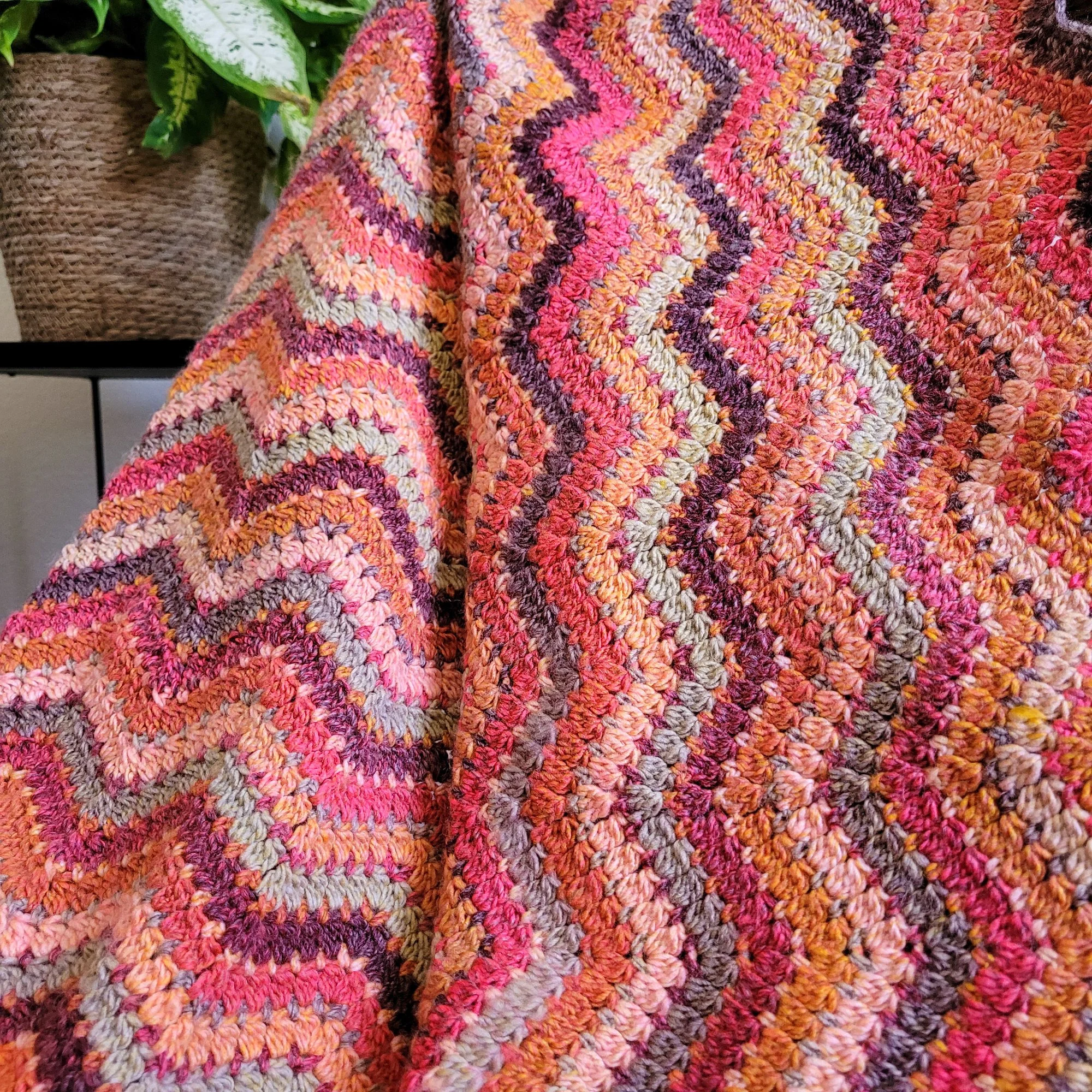 A crochet ripple blanket with wavy stripes in shades of red, orange, brown, and pale green is draped over a chair. A leafy green plant on a metal stand sits behind the blanket to the left.