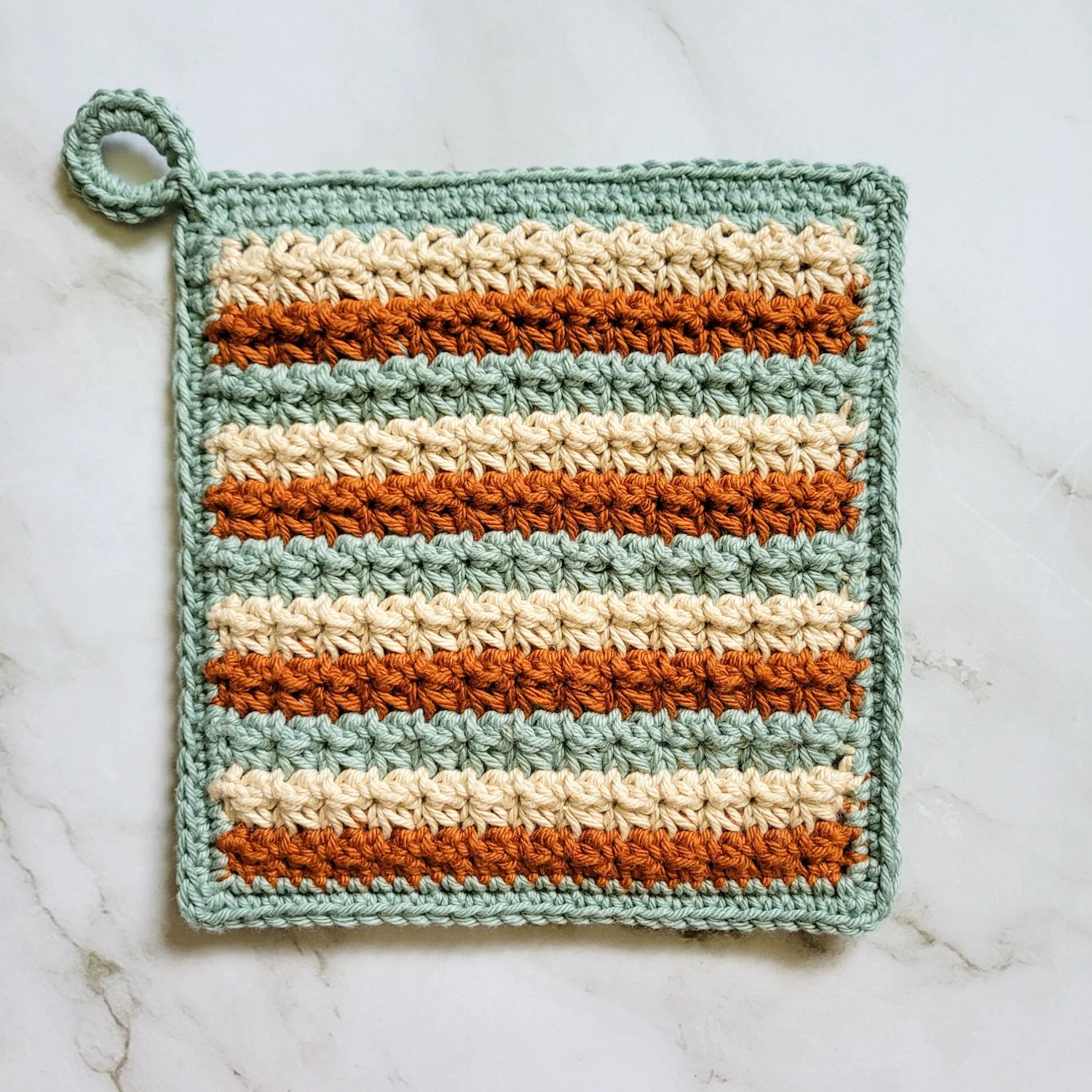 Three-color Classic Potholder crocheted in alternating rows of rust, beige, and sage green Trinity Stitch. The dense, double-thick fabric features a hanging loop and is displayed on a white marble countertop.