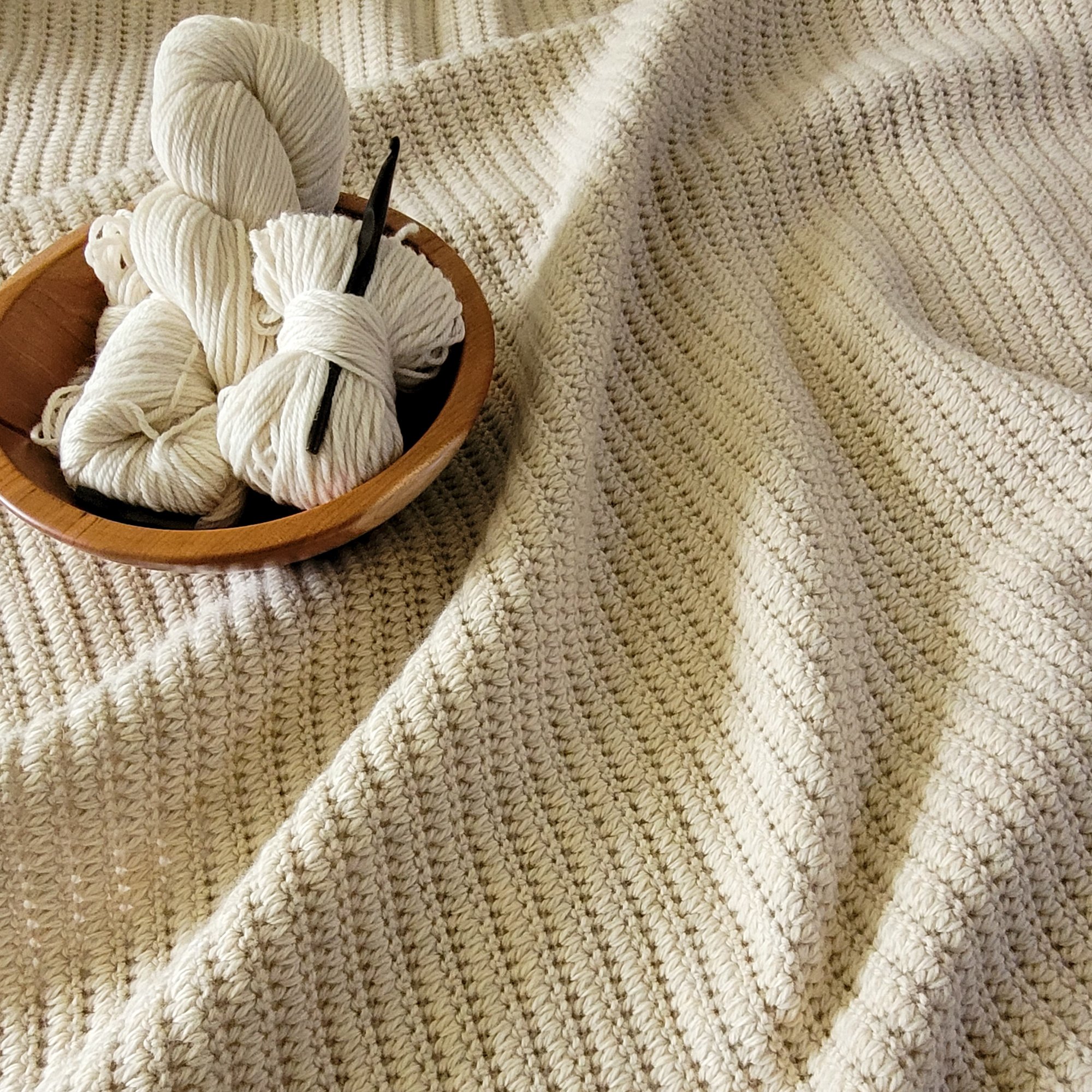 A cream-colored crochet blanket. The interlocking stitch pattern creates a distinct texture. A wooden bowl with balls of cream-colored yarn and a wooden crochet hook sits on the blanket.