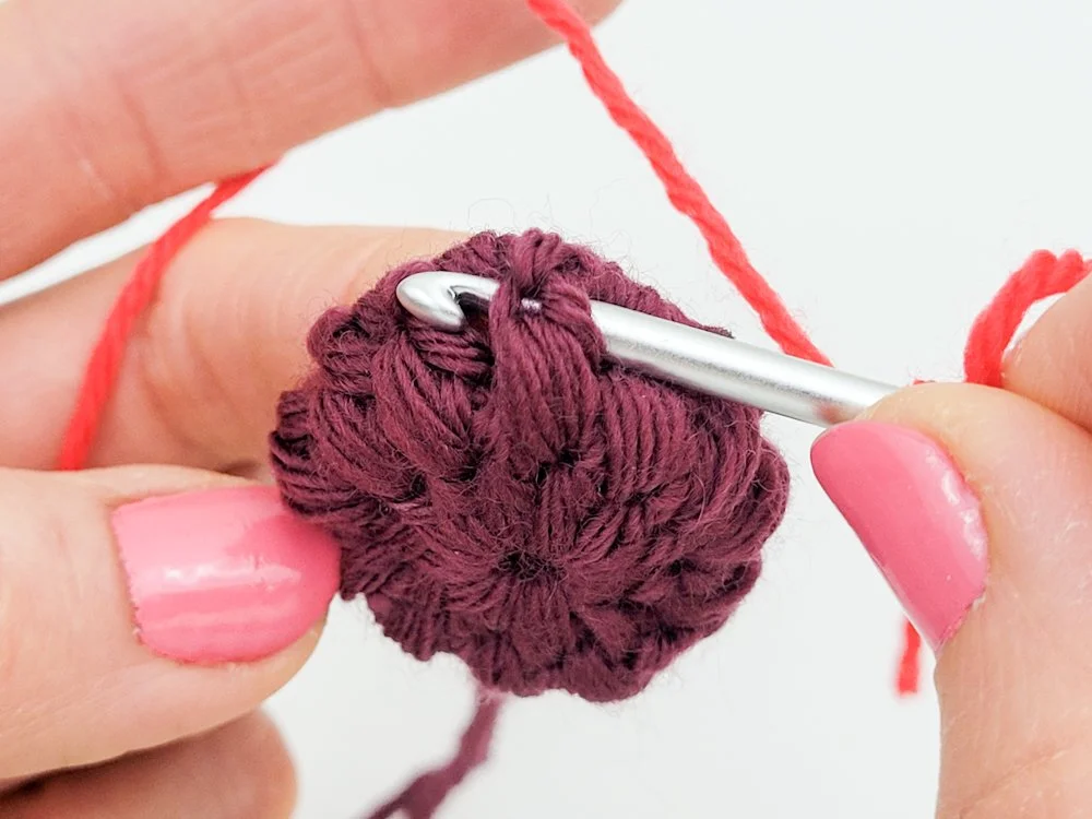 A circle crocheted in dark purple yarn. A crochet hook has been inserted around the post at the top of one of the puff stitch.